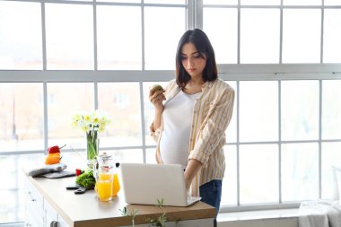 Elmalı hamile bir kadın mutfakta dizüstü bilgisayar kullanıyor.