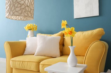 Oturma odasındaki masada minderleri ve çiçek açan narsist çiçekleri olan rahat sarı kanepe.