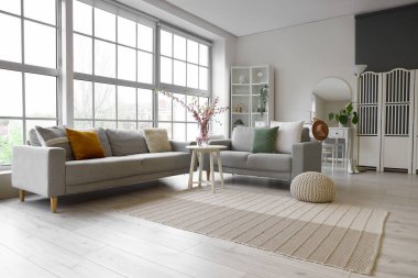 Oturma odasının içinde gri kanepeler ve kahve masasının üzerinde çiçek açan sakura dalları büyük pencerenin yanında.