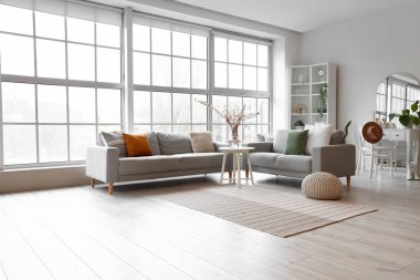 Interior of living room with grey sofas and blooming sakura branches on coffee table near big window clipart