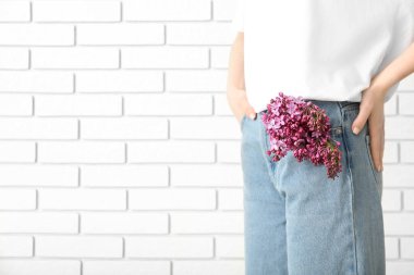 Kotunun cebinde leylak çiçekleri olan beyaz tuğla duvara karşı duran bir kadın.