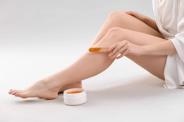 Young woman applying sugaring paste onto legs against light background