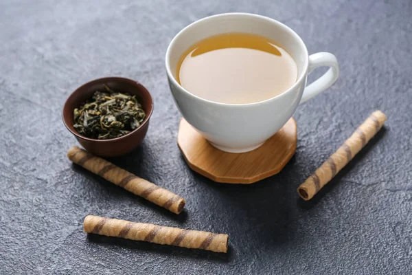 Drink coasters with cup of tea, dry leaves in bowl and wafer rolls on dark table