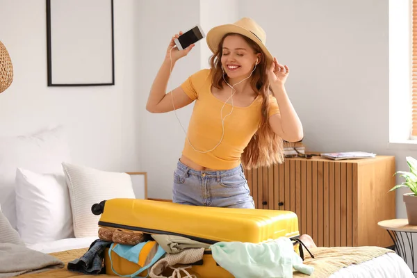 stock image Young woman with earphones and mobile phone packing her clothes for travelling in bedroom