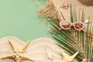 Palmiye yaprağı, kum, deniz kabuğu ve yeşil arka planda plaj aksesuarları olan kompozisyon
