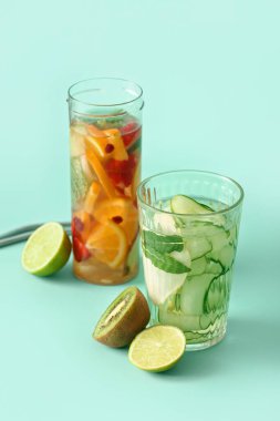 Turkuaz arka planda dilimlenmiş farklı meyvelerle dolu bir bardak ve bir şişe erimiş su.