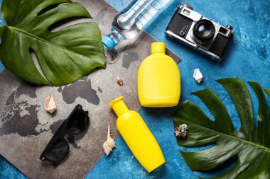 Dünya haritalı kompozisyon, güneş kremi şişeleri, mavi masada tropikal yapraklar ve güneş gözlükleri