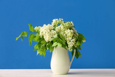 Mavi duvarın yanındaki ahşap masada beyaz leylak vazosu.
