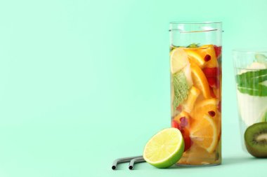 Turkuaz arka planda dilimlenmiş farklı meyvelerle dolu bir bardak ve bir şişe erimiş su.