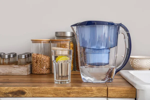 Water filter jug and glass with lemon slice on counter near light wall