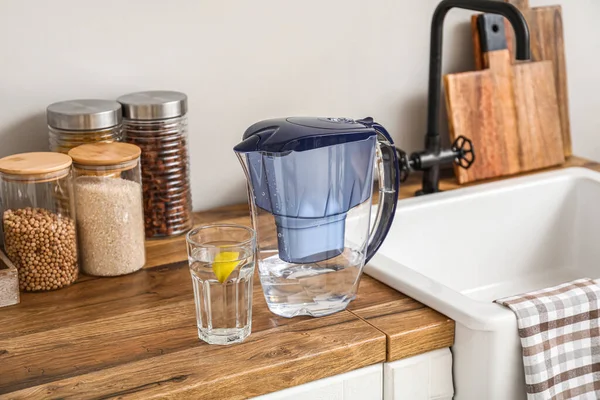 Water filter jug and glass with lemon slice on counter near wall