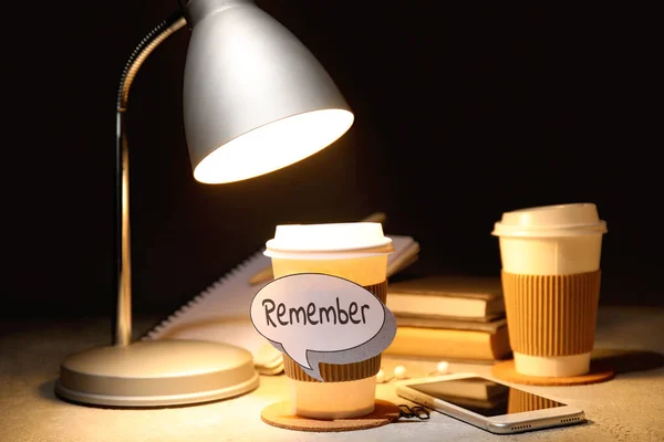 Cup of coffee with reminder, mobile phone and glowing lamp on table, closeup