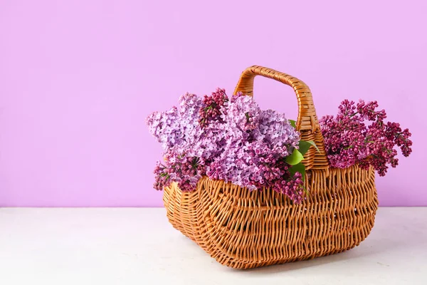 Basket with beautiful lilac flowers on light table against color background