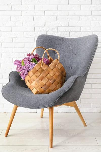 Basket with beautiful lilac flowers on armchair near light brick wall