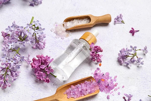 Bottle of lilac essential oil, spoons with sea salt and flowers on light background, closeup