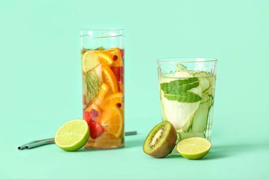 Turkuaz arka planda dilimlenmiş farklı meyvelerle dolu bir bardak ve bir şişe erimiş su.