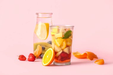 Pembe arka planda dilimlenmiş farklı meyvelerle dolu bir bardak ve bir şişe erimiş su.