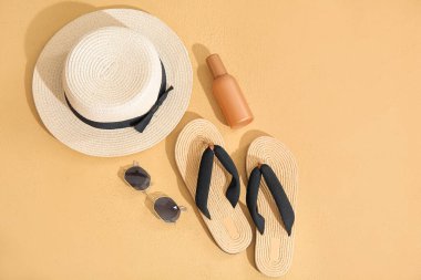 Bir şişe güneş kremi, güneş gözlüğü, arka planda şapka ve parmak arası terlik.