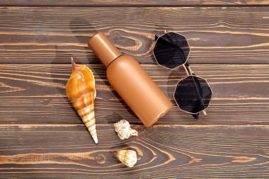 Bir şişe güneş kremi, güneş gözlüğü ve ahşap arka planda deniz kabukları.