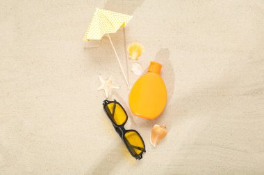 Bir şişe güneş kremi, güneş gözlüğü, şemsiye ve kumdaki deniz kabukları.