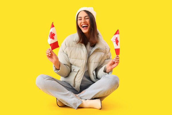 Young woman in warm hat with flags of Canada sitting on yellow background