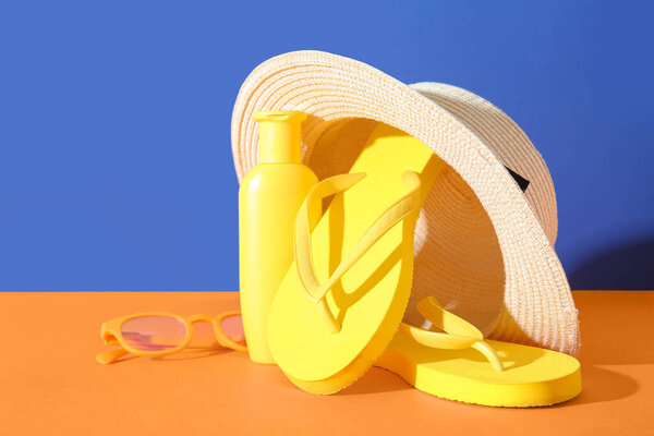 Bottle of sunscreen cream with sunglasses, flip flops and summer hat on orange table near blue wall
