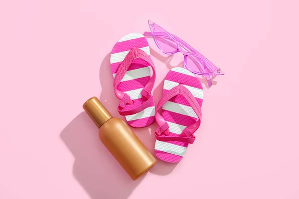Bottle of sunscreen cream with sunglasses and flip flops on pink background