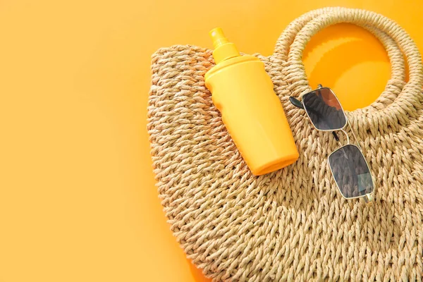 Bottle of sunscreen cream with sunglasses and wicker bag on orange background