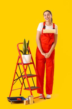 Young woman with ladder, houseplant and paint cans on yellow background