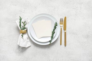 Boş davetiye kartı, çatal bıçak takımı ve beyaz masa üzerinde okaliptüs dallarıyla temiz tabak.