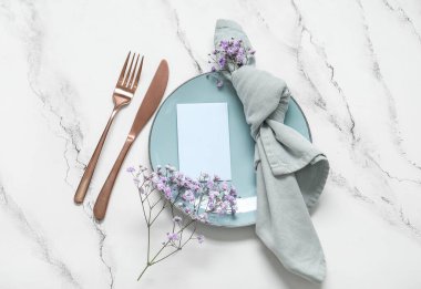 Boş davetiye kartı, çatal bıçak takımı ve beyaz masadaki çigila çiçekleriyle temiz tabak.