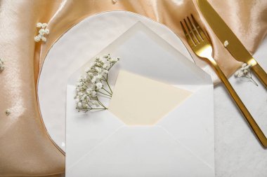 Boş davetiye kartı, çatal bıçak takımı ve beyaz masadaki çigila çiçekleriyle temiz tabak.