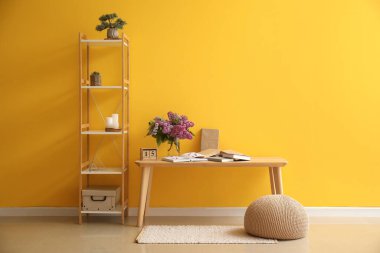 Odanın içinde vazoda leylak çiçekleri, masa ve raf ünitesi var.