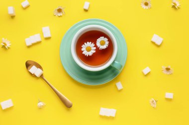 Bir fincan doğal papatya çayı, şeker küpleri ve sarı arka planda çiçekler.