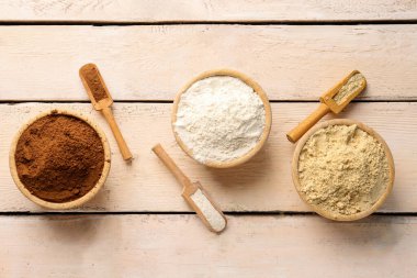 Tahta kepçe ve kâselerle kakao, buğday ve susam unu hafif ahşap arka planda.
