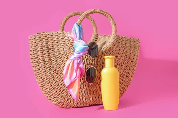 Bottle of sunscreen cream with sunglasses and wicker bag on pink background