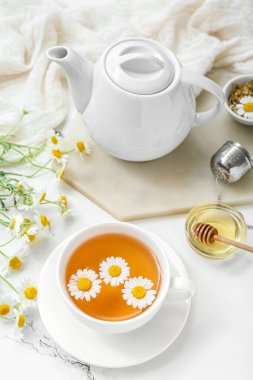 Beyaz mermer arka planda bir fincan sıcak papatya çayı, çaydanlık ve bal.