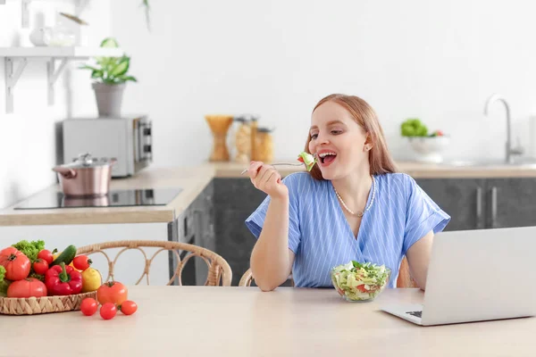 台所で野菜サラダを食べるラップトップを持つ若い女性 — ストック写真