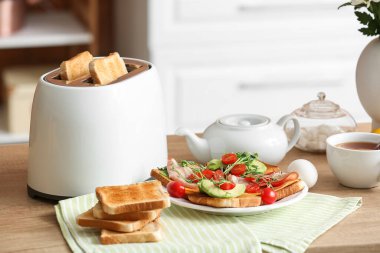 Çıtır ekmek dilimleri, lezzetli sandviçler ve mutfaktaki masada bir fincan çay ile modern tost makinesi.