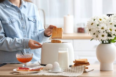 Çağdaş ekmek kızartma makinesinden çıtır ekmek dilimleri alan bir kadın mutfakta kahvaltıyla masada oturuyor.
