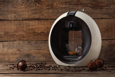 Damlayan espresso, bardak, kapsül ve fasulye ile modern kahve makinesi ahşap masada.