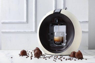 Damlayan espresso, fincan, kapsül kahve makinesi ve beyaz masa üzerinde fasulye.