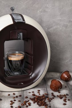 Gri grunge masasında bir fincan espresso, kozalar ve fasulye ile modern kapsül kahve makinesi.