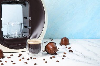 Bir bardak nefis espresso, kapsül kahve makinesi, kapsüller ve fasulyeler beyaz masada.