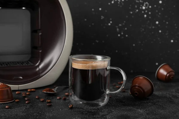 stock image Glass cup of delicious espresso, capsule coffee maker, pods and beans on black grunge table