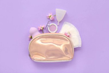 Leylak arka planında regl fincanı, ped ve makyaj ürünleri olan bir kadın makyaj çantası.