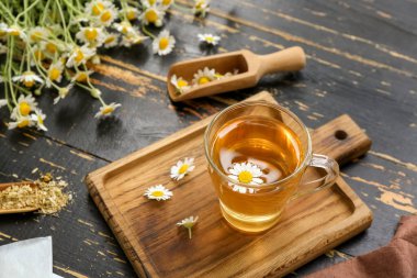 Board with cup of hot chamomile tea and scoops on black wooden background clipart