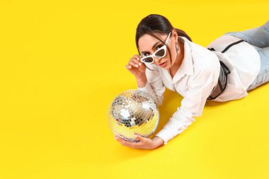 Şık güneş gözlüklü ve sarı arka planda duran disko toplu güzel bir genç kadın.