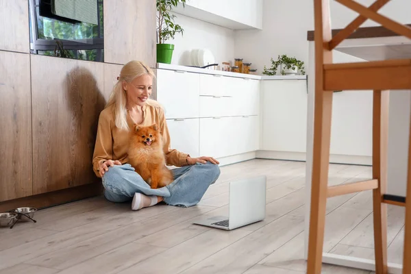 Mature woman with Pomeranian dog and laptop in kitchen