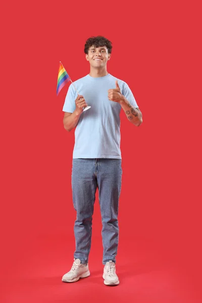 Lgbtの旗を持つ若い男が赤い背景に親指アップを示す — ストック写真